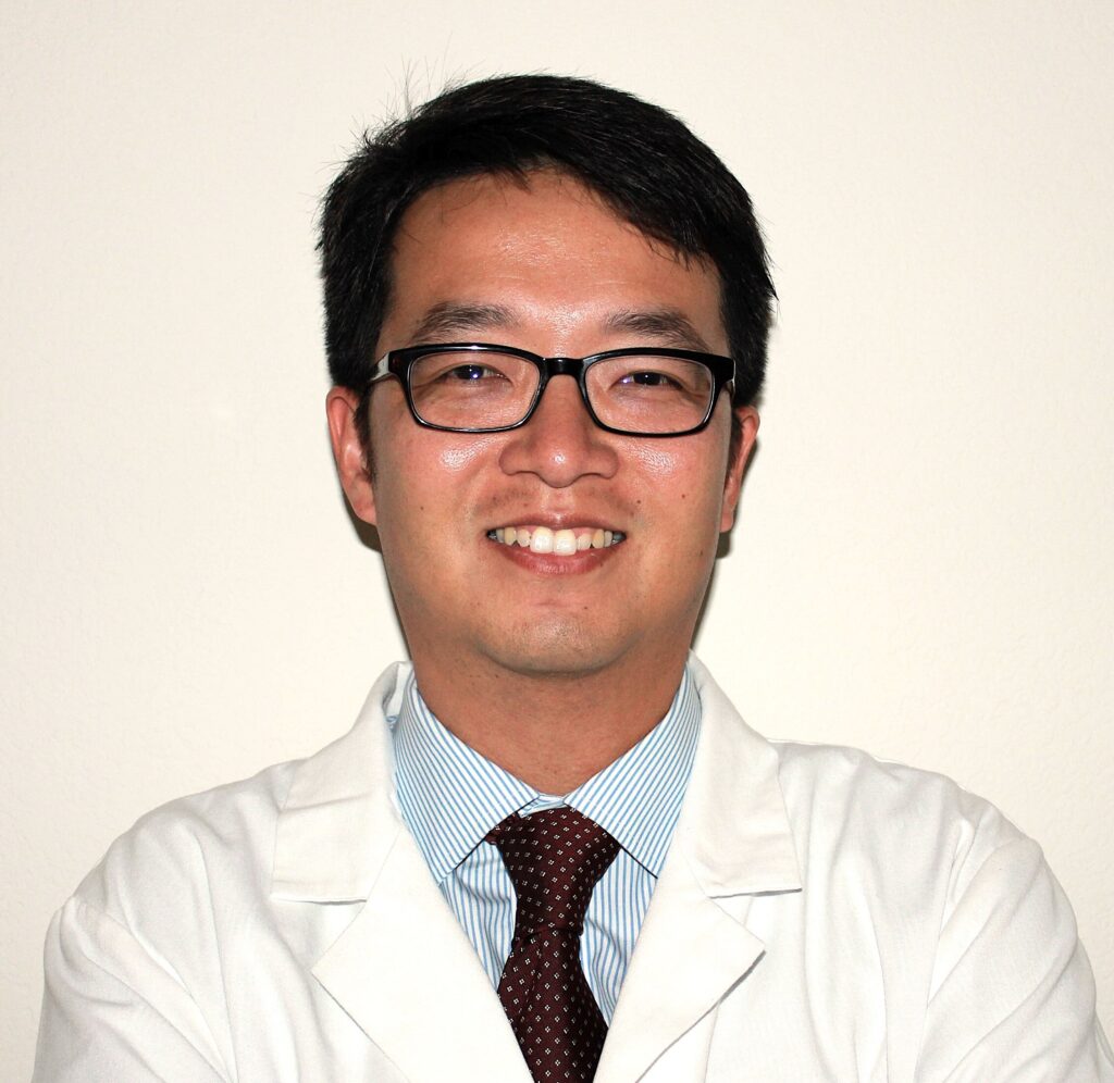 Close-up of a man wearing glasses, a white lab coat, and a blue striped shirt with a maroon tie, smiling at the camera.