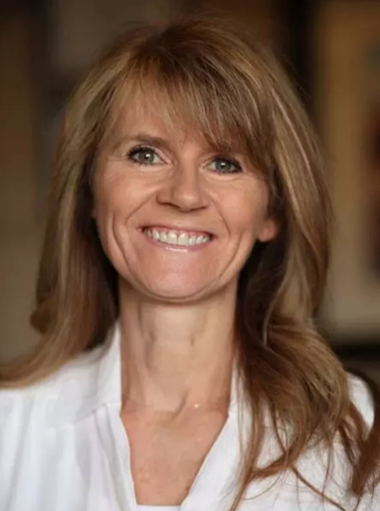 Smiling woman with shoulder-length light brown hair wearing a white blouse, neutral indoor background.